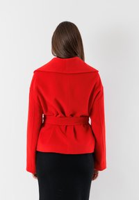 Giacca in misto lana rossa con ampio colletto, vita con cintura e maniche lunghe, caratterizzata da una vestibilità su misura e dettaglio plissettato sul retro.