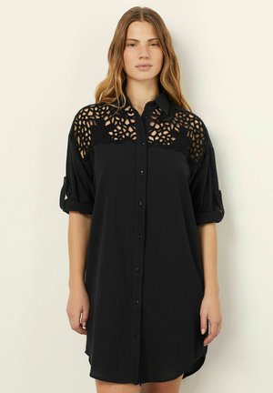 Vestido negro de camisa con mangas cortas enrolladas, cierre de botones en la parte delantera y un intrincado detalle de ganchillo en la zona de los hombros. Tejido suave y texturizado.