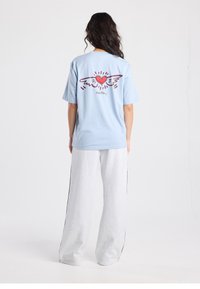 Lichtblauw katoenen T-shirt met korte mouwen en een rood hart met vleugelafbeelding op de achterkant. Gecombineerd met grijze joggingbroek.