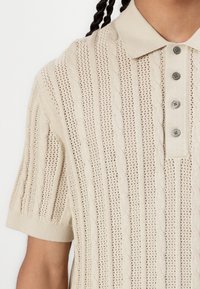 Beige, rövid ujjú polo póló, amely texturált bordás kötésmintával és kábelmintákkal rendelkezik, valamint három gombos záródással. Könnyű és légáteresztő anyag.