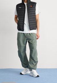 Czarna pikowana kamizelka na białej koszulce, zestawiona z luźnymi zielonymi spodniami cargo i białymi sneakersami. Na kamizelce i koszulce widoczne charakterystyczne logo.