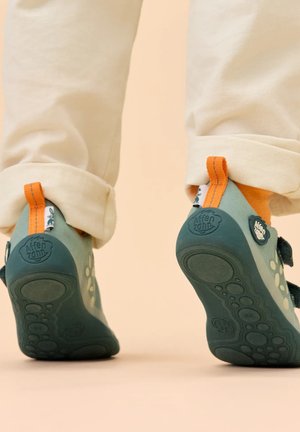 Kind trägt beige Hose und grüne Sneakers mit orangefarbenen Zuglaschen, läuft auf einem glatten beigen Boden, zeigt Schuhsohlen und Absätze.