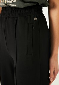 Zwarte tapered broek van zachte stof met een elastische tailleband, een ritszak en een klein zilveren logodetail.