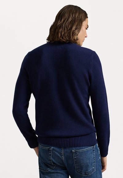 Pulover tricotat bleumarin cu mâneci lungi, margine ribbed și design fără cusături. Purtați cu blugi de culoare albastru deschis, evidențiind țesătura texturată din spate.