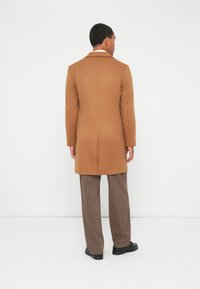 Uomo che indossa un lungo cappotto color cammello, pantaloni a strisce marroni e scarpe nere, in piedi di fronte a un muro bianco con le mani rilassate lungo i fianchi.