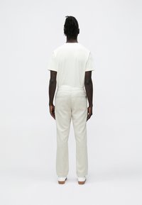 Calças de cor clara, corte solto, com cós plano e dois bolsos traseiros, combinadas com uma t-shirt branca de mangas curtas.