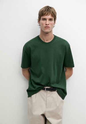 Junger Mann mit kurzen braunen Haaren, der ein dunkelgrünes kurzärmeliges Hemd trägt, das in beige Hose mit schwarzem Gürtel gesteckt ist, steht vor einer weißen Wand.