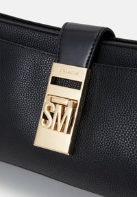 Borsa a mano in pelle nera con finitura testurizzata, una placca dorata con le iniziali "SM" e una chiusura decorativa.