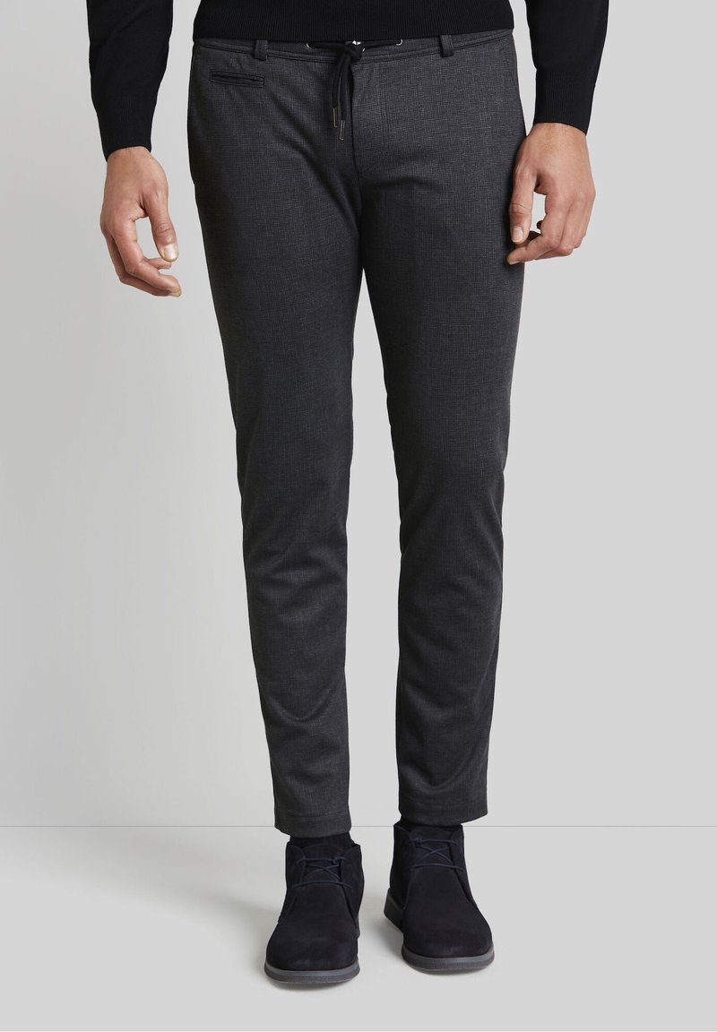 Homme portant un pantalon gris foncé ajusté, un pull noir, des chaussettes noires et des chaussures en daim bleu foncé à lacets, debout sur un sol gris.