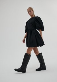 Kobieta idąca w czarnej mini sukience z bufiastymi rękawami, w połączeniu z czarnymi, skórzanymi butami do kolan, na jednolitym tle.