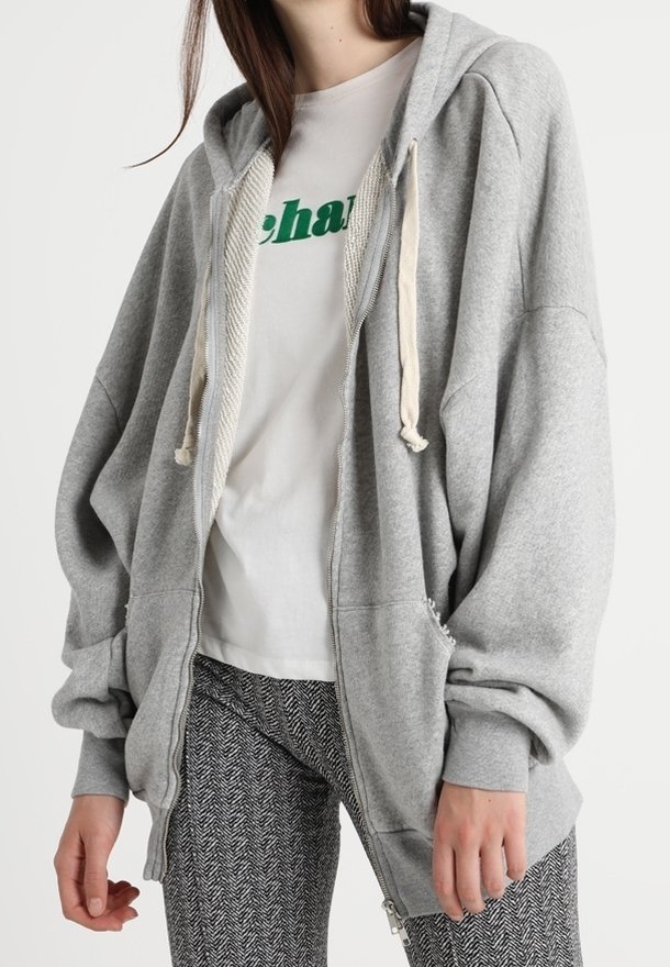 Femme portant un sweat à capuche gris oversize zippé, un t-shirt blanc avec un texte vert et un pantalon à motifs noir et blanc.