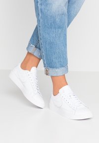 White leather sneakers with a low profile, rounded toe, and lace-up design. Textured detailing and logo on the side. Worn with cuffed denim jeans.