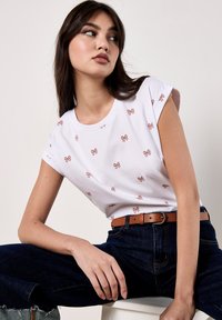 Una joven está sentada con el codo en la rodilla, vistiendo una camiseta blanca con patrones de lazo rosa y jeans azul oscuro, mirando pensativa hacia un lado.