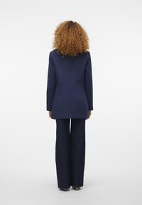Blazer bleumarin cu o croială ajustată, având umeri structurați și o textură fină, asortat cu pantaloni din denim întunecat, cu picior larg.