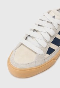 Tenisice s gornjim dijelom od svijetlosive ante, bijelim vezicama, mornarskim trakama i gumiranim potplatima; okrugli vrh i podstavljen ovratnik.