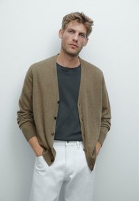 Cardigan marrone infilato sopra una t-shirt scura, abbinato a pantaloni bianchi; texture morbida e vestibilità rilassata, con polsini e orlo a coste.