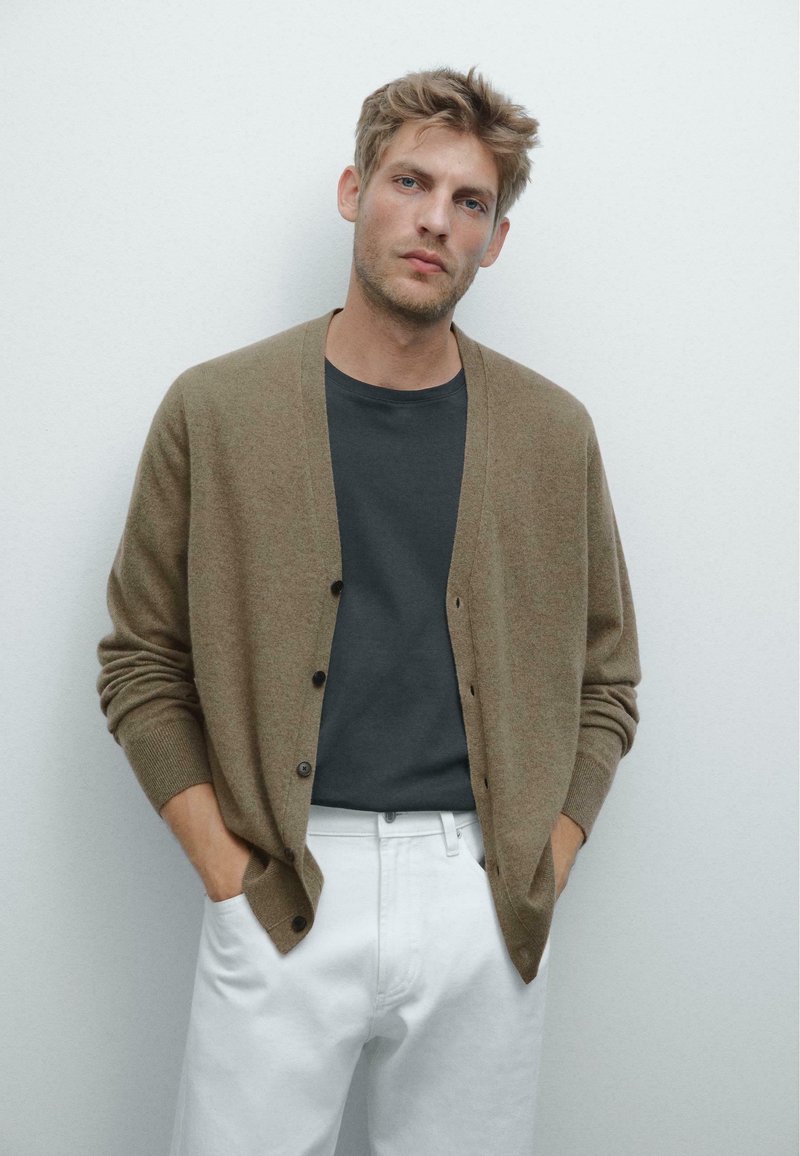 Cardigan marrone infilato sopra una t-shirt scura, abbinato a pantaloni bianchi; texture morbida e vestibilità rilassata, con polsini e orlo a coste.