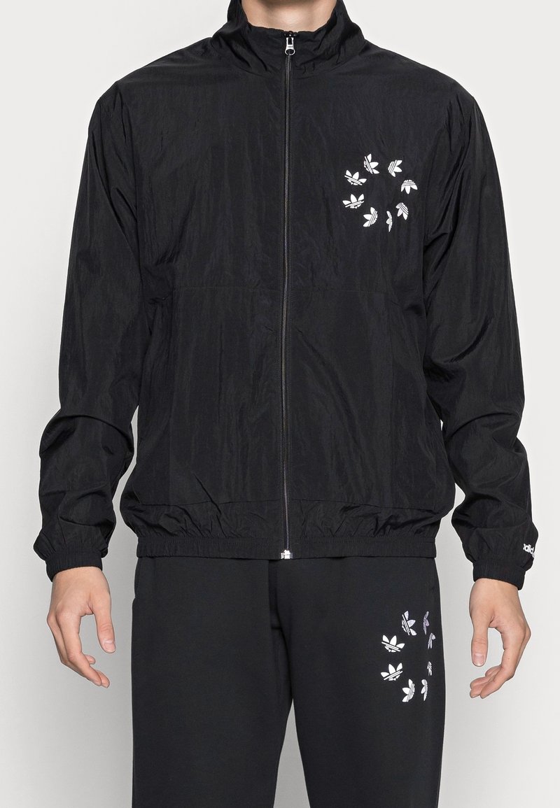 Schwarze leichte Jacke mit hohem Kragen und Reißverschluss vorne. Verfügt über weiße Logo-Akzente und elastische Bündchen. Kombiniert mit schwarzen Hosen mit ähnlichen Details.