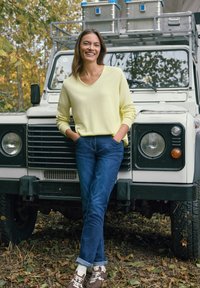 Femme en pull jaune et jean bleu appuyée contre un véhicule tout-terrain blanc dans une zone boisée avec des feuilles tombées.