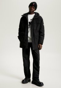 Parka negra aislante con múltiples bolsillos, cremallera y capucha con cordón. Usada sobre un suéter blanco y combinada con pantalones y botas negras.