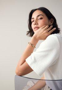 Gold chain bracelet on wrist, with a smooth texture. White short-sleeve shirt contrasts against the skin. Simple, clean design.