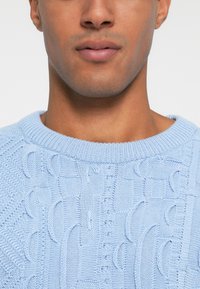 Ανοιχτό μπλε πλεκτό πουλόβερ με υφή, ριμπ μανίκι και κοντά μανίκια, που αναδεικνύει πολύπλοκες λεπτομέρειες ραφής.