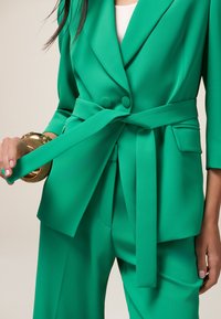Groen op maat gemaakt kostuum met een ceintuur in de taille, voorzien van grote knopen en een geweven stof. De outfit heeft een brede revers en strakke broeken.