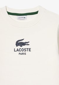 Kremfarget hettegenser med rund hals, med en mørkeblå krokodillogo og "LACOSTE PARIS" tekst under. Grønn innvendig kant.