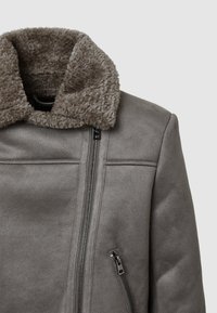 Giacca in finta pelle scamosciata grigia con un colletto in pelliccia beige soffice, dotata di una zip frontale e di una tasca laterale con zip. Texture liscia e design strutturato.