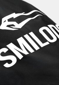 Sort tekstureret stof med stor hvid delvis tekst "SMILODS" og en stiliseret hvid håndgrafik over teksten.