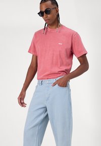 Ροζ βαμβακερό t-shirt με εκτύπωση "obey", συνδυασμένο με ανοιχτό μπλε τζιν παντελόνι, που διαθέτει άνετη εφαρμογή και πλαϊνές τσέπες. Φοριούνται μαύρα γυαλιά ηλίου.