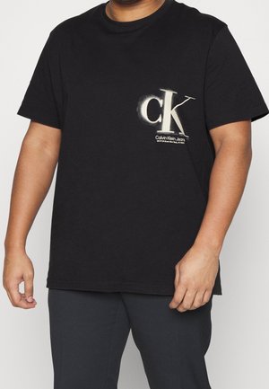 Osoba nosząca czarną koszulkę Calvin Klein Jeans z białym logo "ck" i tekstem na lewej piersi, w zestawieniu z ciemnymi spodniami.