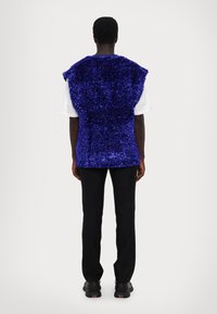 Purpurinė, blizganti be rankovių liemenė, laisvai krentanti, dėvima ant baltos marškinėlių, derinama su juodomis kelnėmis ir tamsiais sportbačiais.