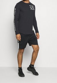 Mies, który ma na sobie czarną koszulę z długim rękawem z białym napisem, czarne szorty z białymi paskami, czarne skarpetki Nike i czarne buty sportowe.