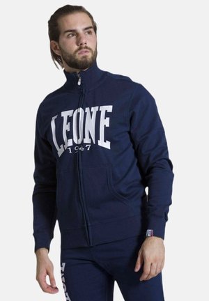 Uomo che indossa una giacca blu navy con zip e scritta "LEONE 1947" e pantaloni abbinati, guarda leggermente a sinistra.