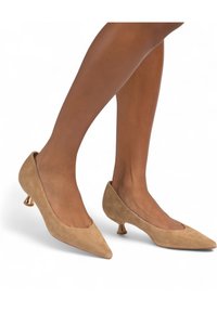 Beige suède hoge hakken met een puntige neus en metalen gouden accenten op een lage blokhak. Gladde textuur met een matte afwerking.