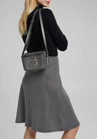 Šedá crossbody taška s texturovaným povrchem, vyznačující se výrazným logem. V kombinaci s černým topem a sukní s houndstooth vzorem.