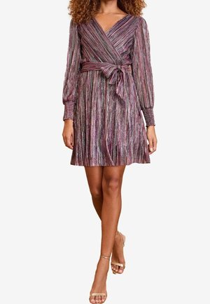 Kobieta w długim rękawie, sukience o długości do kolan, kopertowej, z metalicznymi pionowymi paskami i przewiązanym paskiem w talii, w połączeniu z sandałkami na paski.