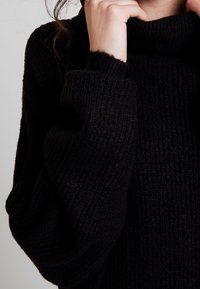 Mustassa neulotussa turtleneckissa on tekstuuripinta. Lievät hihat ja korkea kaulus luovat rentoa siluettia.