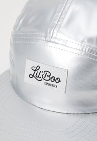 Berretto argentato realizzato in materiale lucido, con una visiera cucita, superficie testurizzata e un'etichetta bianca ben visibile con la scritta "Lil' Boo Copenhagen."
