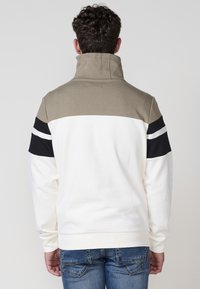 Mikina s okrúhlym výstrihom s vysokým golierom, béžová horná časť, biele telo a čierne pruhy na pleciach. Mäkko textúrovaná.