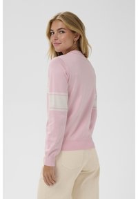 Joven mujer con cabello rubio que lleva pendientes de aro dorados, un suéter rosa con rayas blancas en las mangas y pantalones beige, mirando por encima de su hombro.