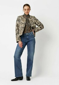 Texturierte goldene und schwarze gemusterte Jacke, hoher Kragen, figurbetont über einem braunen Oberteil, kombiniert mit blauen geraden Jeans und schwarzen Ankle Boots.