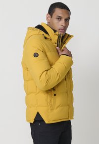 Giacca imbottita gialla con cappuccio, chiusura a zip e patch con logo sulla manica. Tessuto testurizzato con motivo trapuntato e tasche laterali.