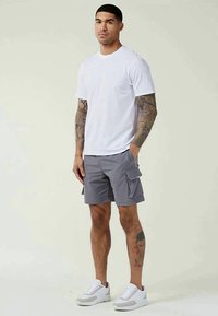Man with tattoos wearing a white t-shirt, gray cargo shorts, and white sneakers, standing with one hand in pocket against plain background.