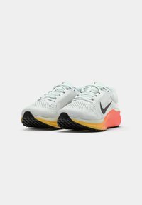 Zapatillas de correr gris claro con parte superior de malla, logo Swoosh negro, suela naranja y amarilla, agarre texturizado y diseño con cordones.