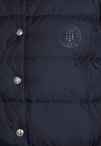 Marineblå pufferjakke med en blank finish, quiltet tekstur, metaltrykknapper og et diskret logo-print med året 1985.