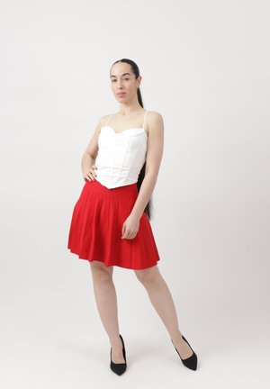 Kvinde med langt mørkt hår iført en hvid korsettop, rød plisseret nederdel og sorte hæle, stående mod en ensfarvet hvid baggrund.