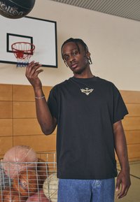 Sort bomuld T-shirt med et centralt gyldent emblem, parret med blå denim jeans. Baggrunden inkluderer en basketballkurv og en trådkurv med bolde.