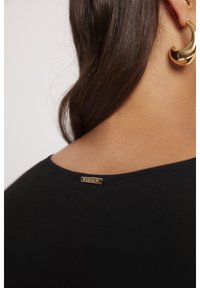 Czarna koszulka z okrągłym dekoltem, z złotym metkiem na plecach. Gładka tekstura, zbliżenie pokazuje kolory włosów i skóry.